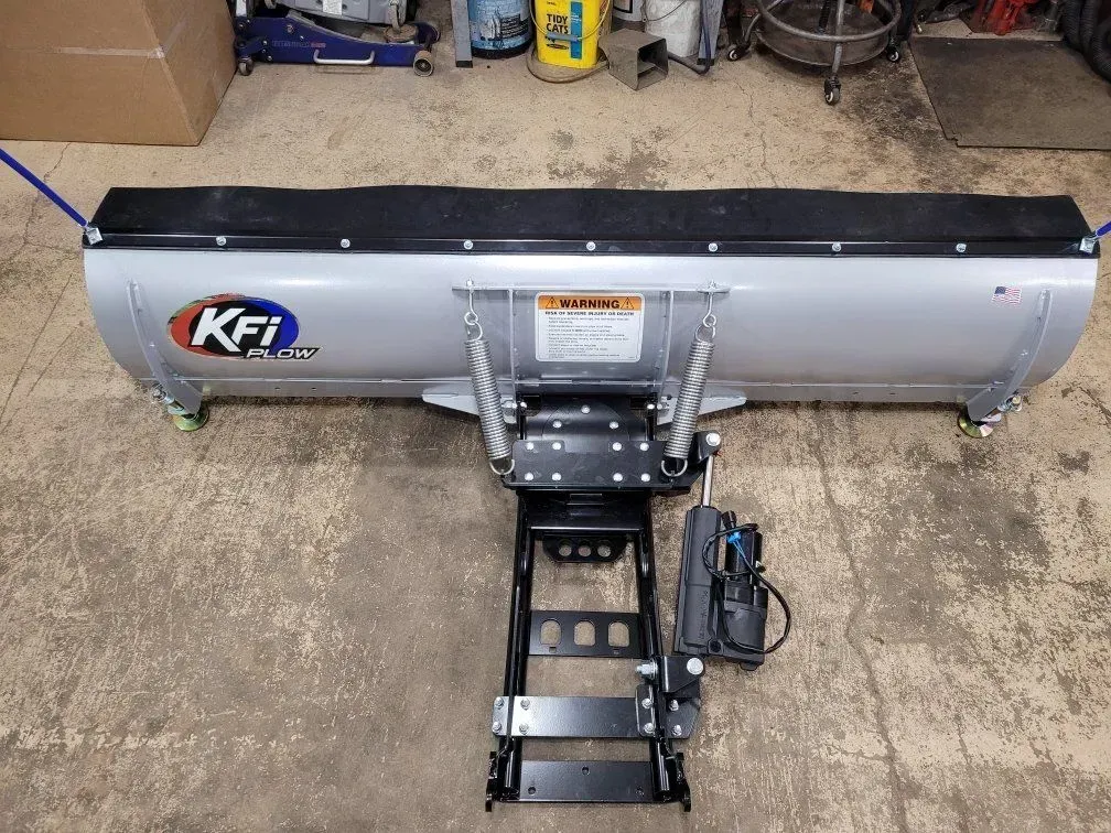 Silver KFI snow plow on a black mounting frame, sitting on a concrete floor.